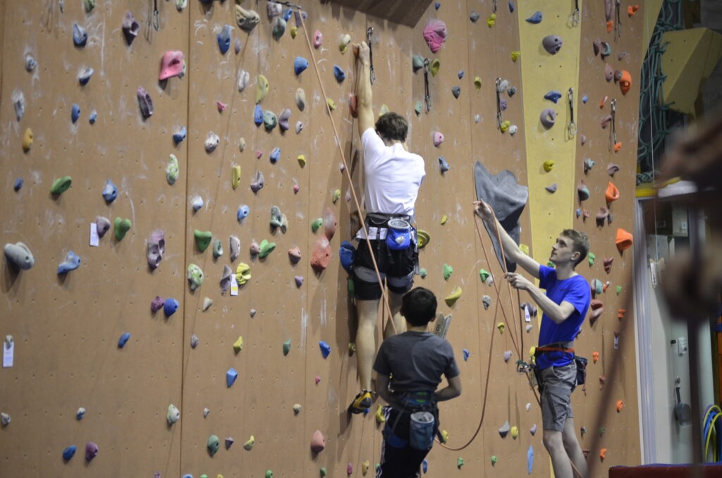 2 jeunes assurent des grimpeurs qui escaladent une structure artificielle d'escalade.
